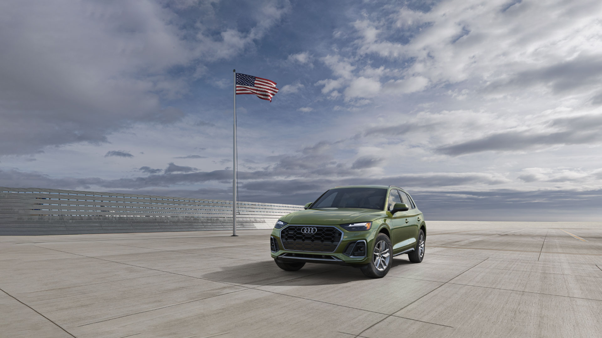 An Audi A6 allroad vehicle parked on an open tarmac with a American flag waving on a flagpole.